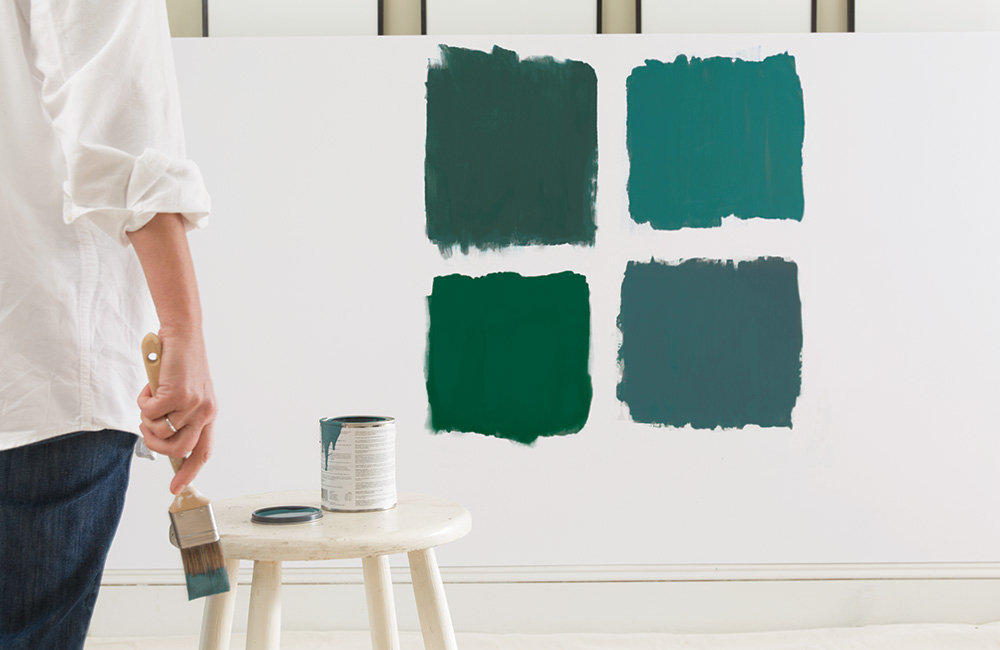 Interior vignette featuring woman painting swatches on the wall with stool, paint can and brush.Color A (Upper left): Hunter Green 2041-10.Color B (Upper right): Teal Ocean 2049-30.Color C (Lower right): True Green 2042-10.Color D (Lower left): Seaweed 2035-10.