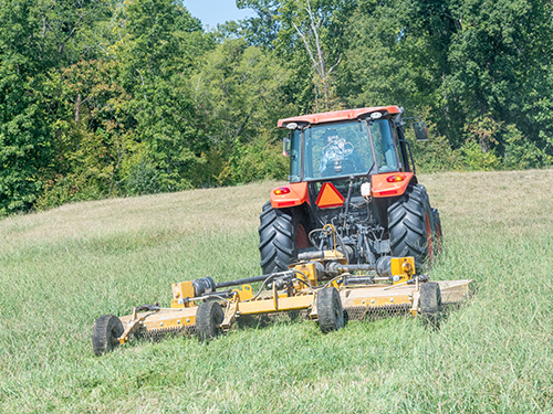 A BushWhacker cutter being pulled by a tractor through a field.