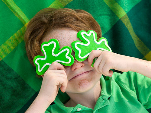 A kid holding three leaf clover cookies over his eyes while he wears a green polo and is laying on a green blanket