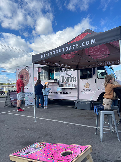 A donut food truck with a line of people in front of it. They have donut themed corn hole boards sitting on the ground out front.